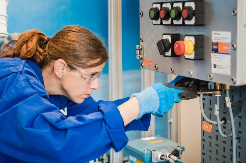 A woman in safety goggles and a blue lab coat uses a wrench to adjust components on an industrial control panel.