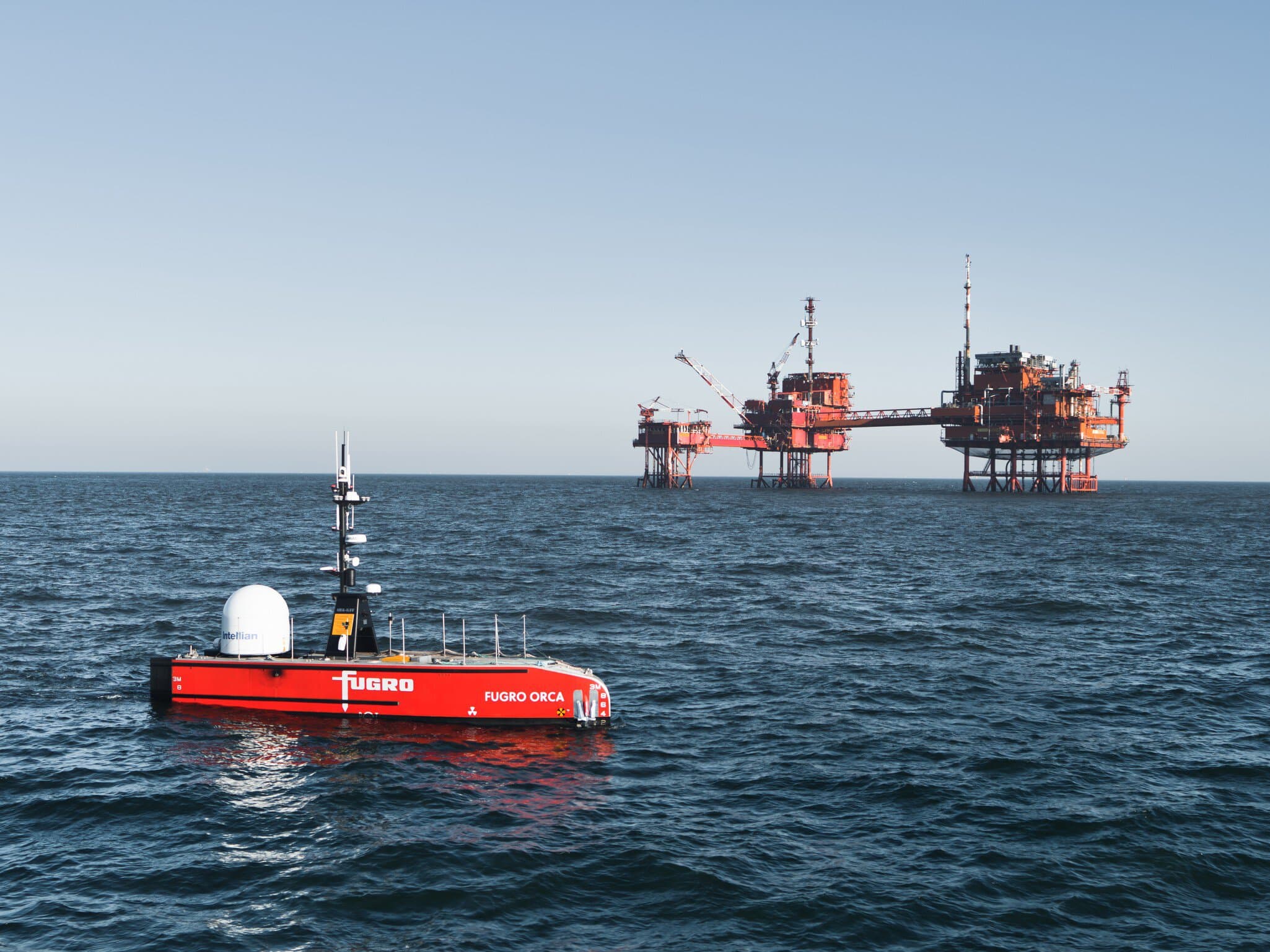 A red unmanned surface vehicle labeled "Fugro Orca" floats in the ocean near an offshore oil platform under a clear sky.